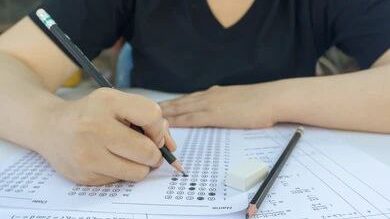 एक काले T-shirt में लड़की हाथ में पेंसिल को पकड़ी हुई परीक्षा दे रही है। Exam MCQ types का है। और पेपर पर दूसरी एक pencil और eraser रखी गई है।